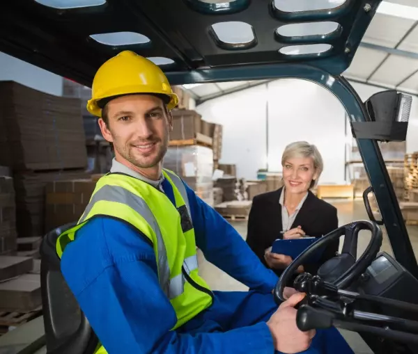 Man met helm en fluovest op heftruck met een vrouw in maatpak op de achtergrond